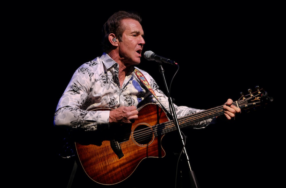 Dennis Quaid in white shirt playing an acoustic guitar and singing in a mic