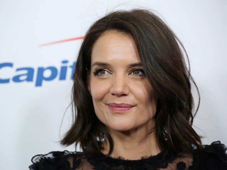 Katie Holmes smiles in a black dress against a white background