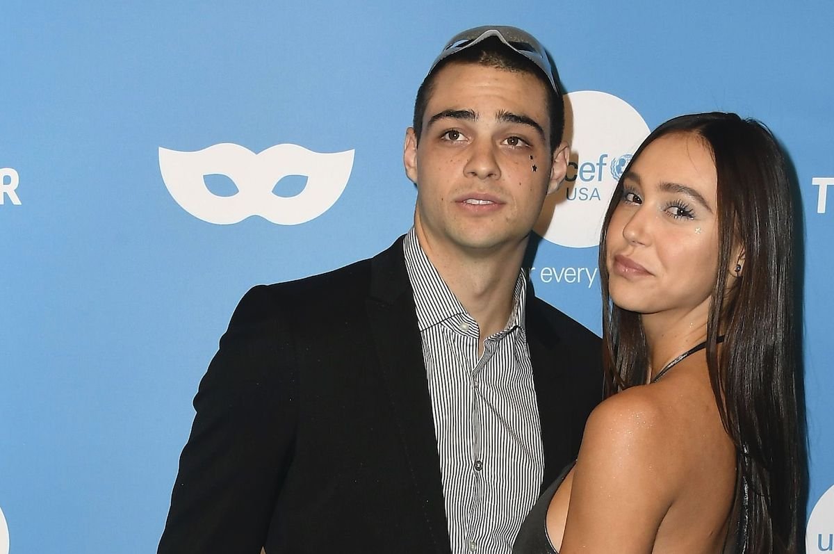 Noah Centineo looks off camera in a black and grey suit with his arm around Alexis Ren in a shiny si