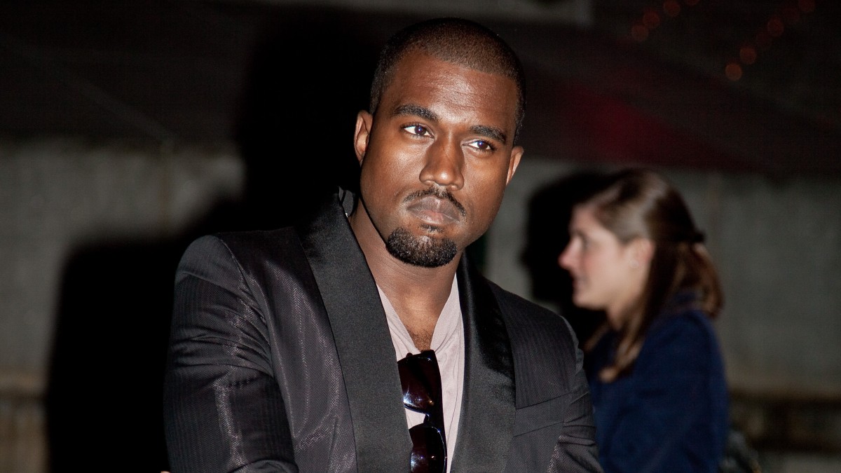 Kanye West wears a dark suit jacket over a t shirt and crosses his arms
