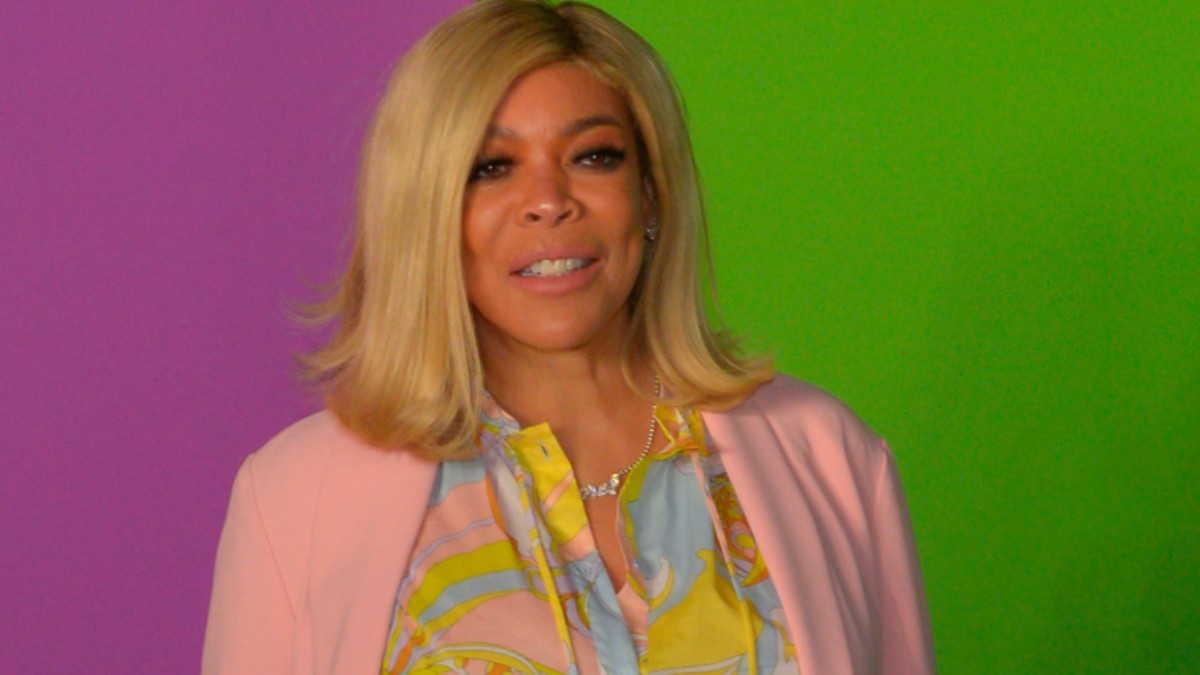 Wendy Williams stands against a purple and green background while wearing a pink top