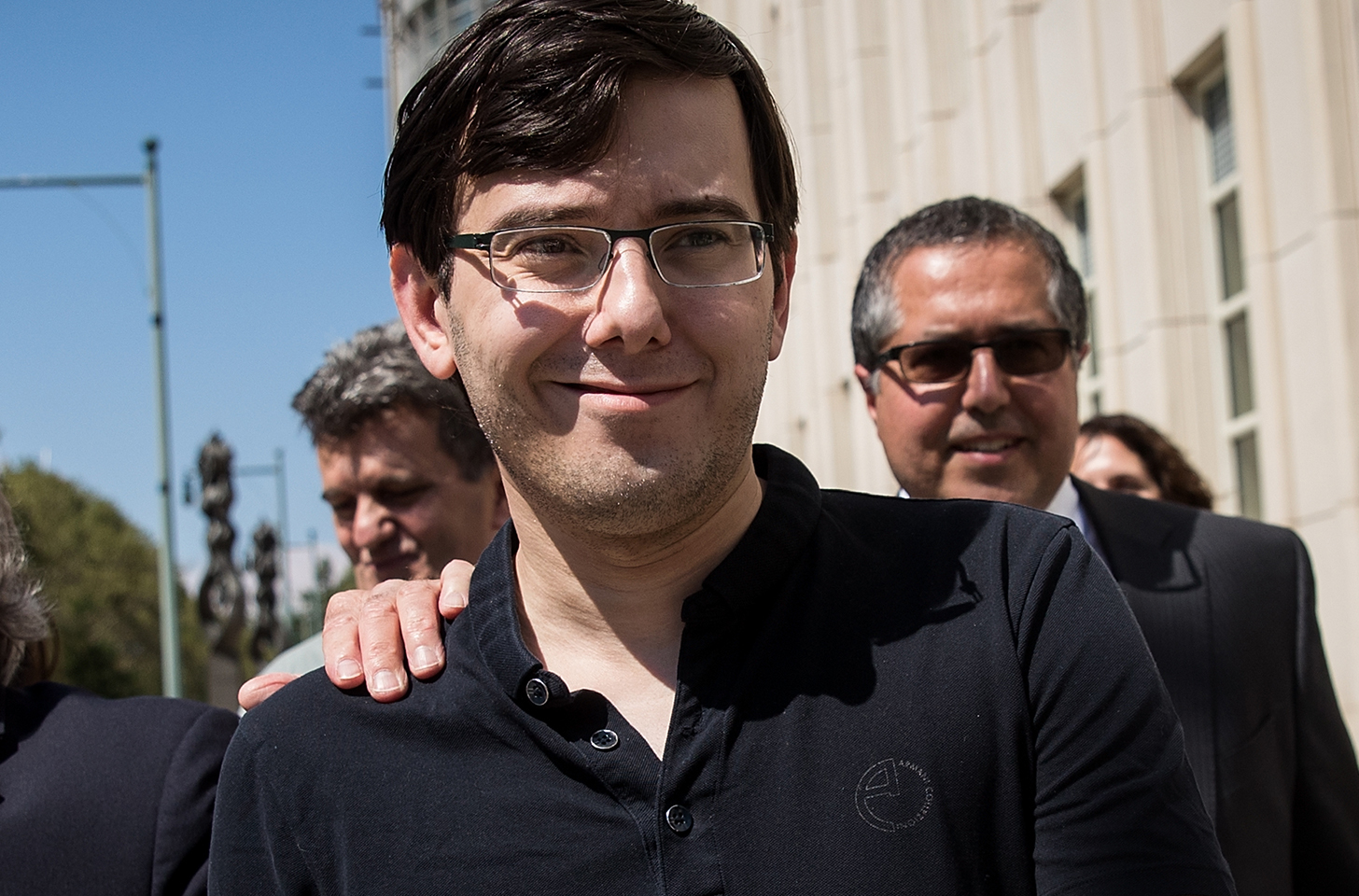 Martin Shkreli in a black polo shirt, walking out of court, smiling.