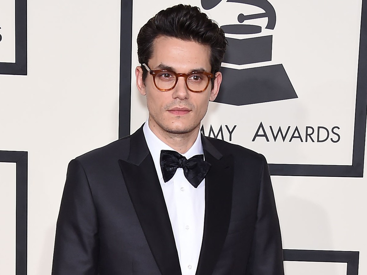 John Mayer poses in black suit and bowtie/glasses against white backdrop