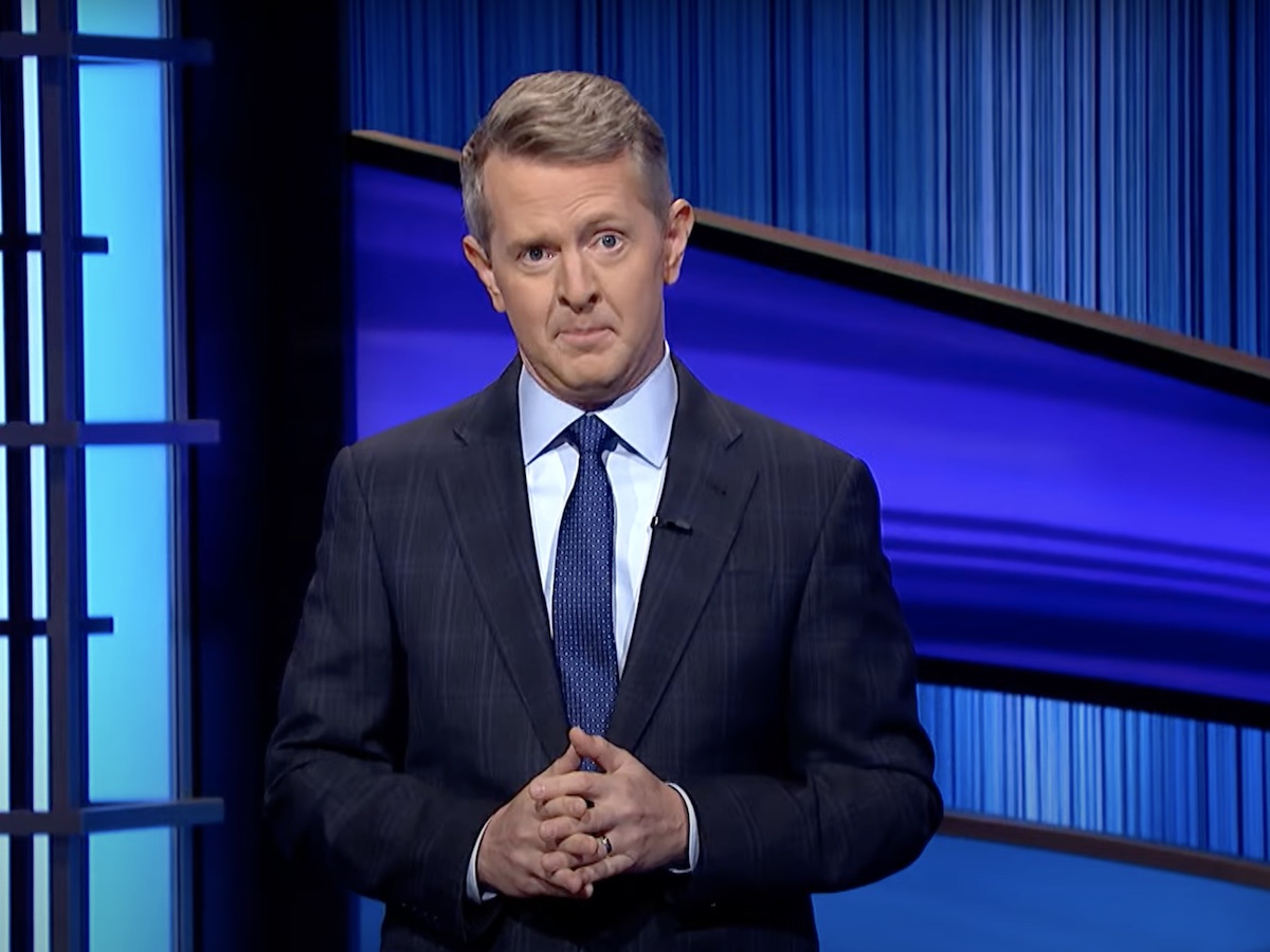 screenshot of Ken Jennings introducing Jeopardy in a navy suit and tie with an eyebrow raised and his hands clasped in front of him